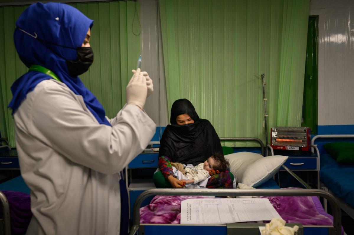 Una mujer afgana sentada en su cama con su nieta de un mes y medio, que padece desnutrición aguda grave y neumonía, mientras una enfermera se prepara para administrar a la niña medicación intravenosa en una sala de hospitalización de la unidad de alimentación terapéutica de Acción contra el Hambre